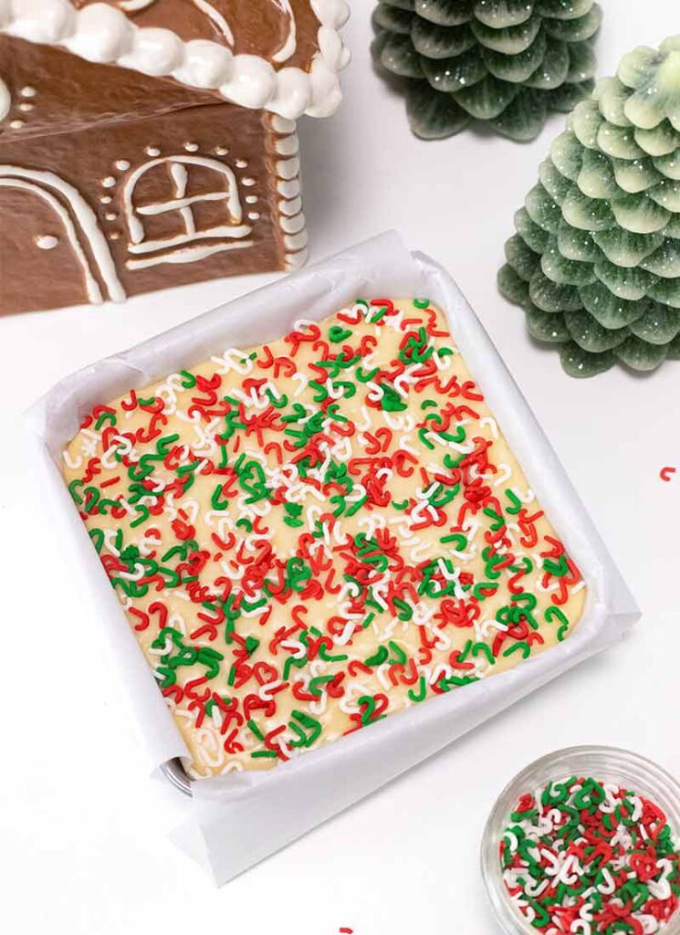 A square baking dish filled with uncut vanilla fudge, topped with red, white and green sprinkles. The dish is lined with parchment paper. A gingerbread house, a jar of sprinkles and pine tree decorations are around the fudge.
