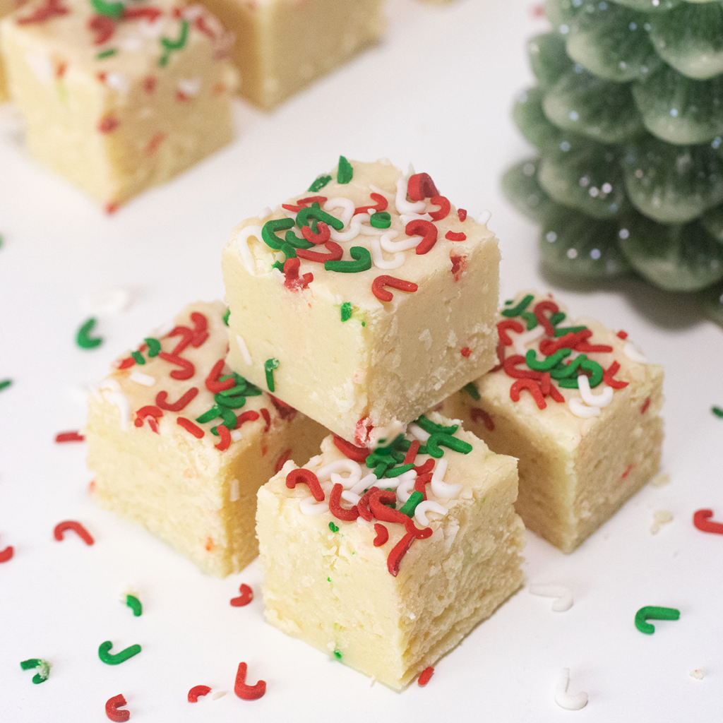 Cubes of white fudge are stacked on a white surface. The fudge is decorated with red, white and green sprinkles.