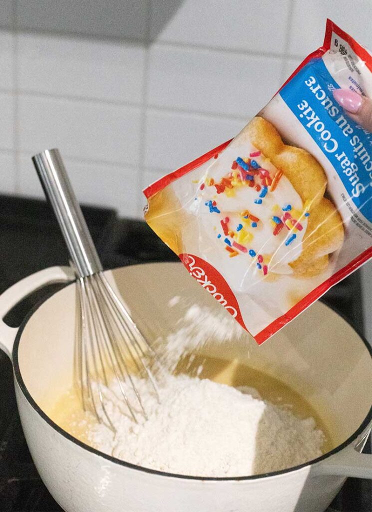 A bag of sugar cookie mix is being poured into a pot on the stove. A wire whisk is resting in the pot.