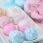 A white, rectangular plate topped with 8 small balls of cotton candy, in shades of blue, pink and orange. More blue and pink cotton candy sits behind the plate, to the right.