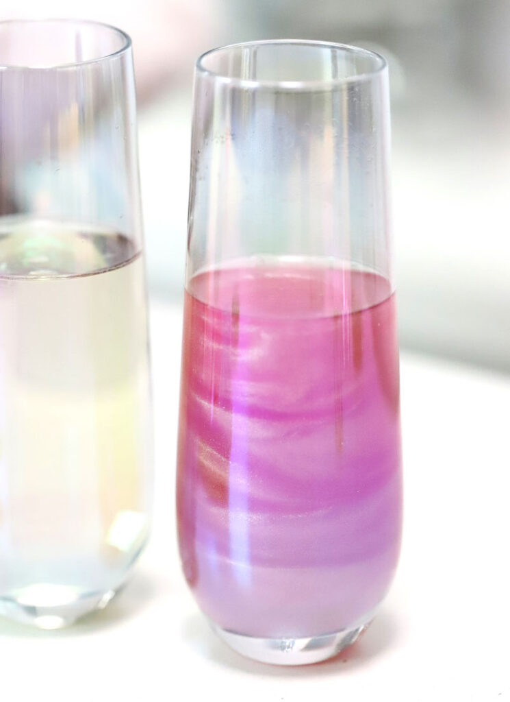 Two stemless champagne flutes are on a white surface. One is filled with clear champagne, the other is filled with shimmery pink champagne.