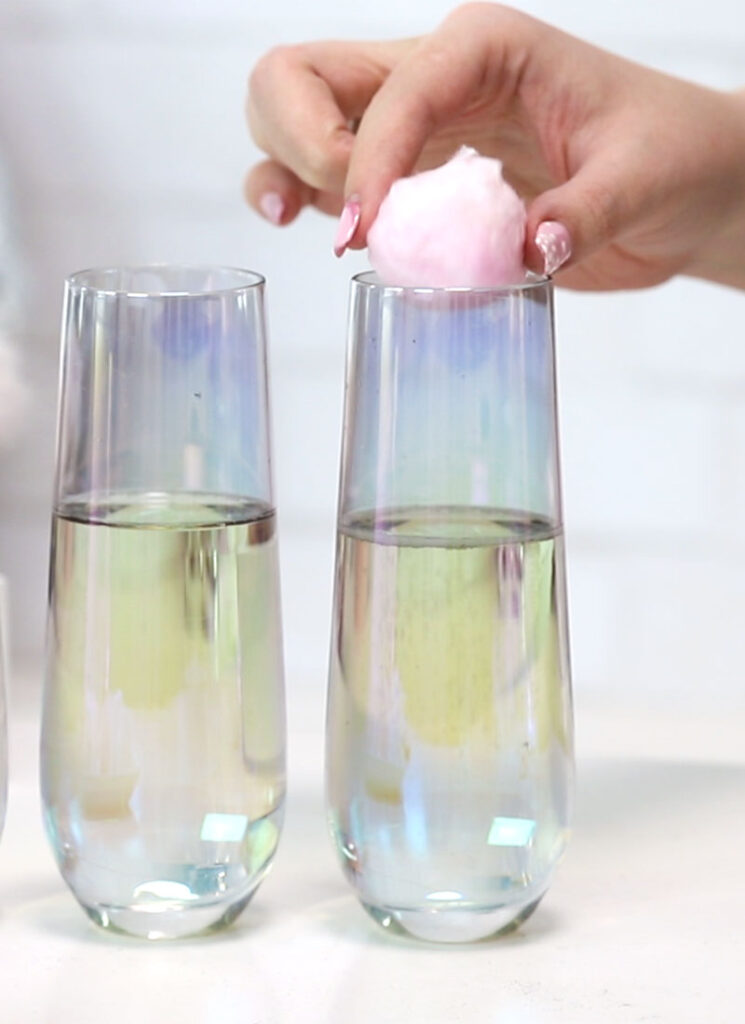 Two stemless champagne flutes are on a white surface. Both are filled with champagne. A hand is lowering a ball of cotton candy into one of the glasses.