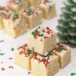 Cubes of white fudge are stacked on a white surface. The fudge is decorated with red, white and green sprinkles. A pine tree decoration is in the background.