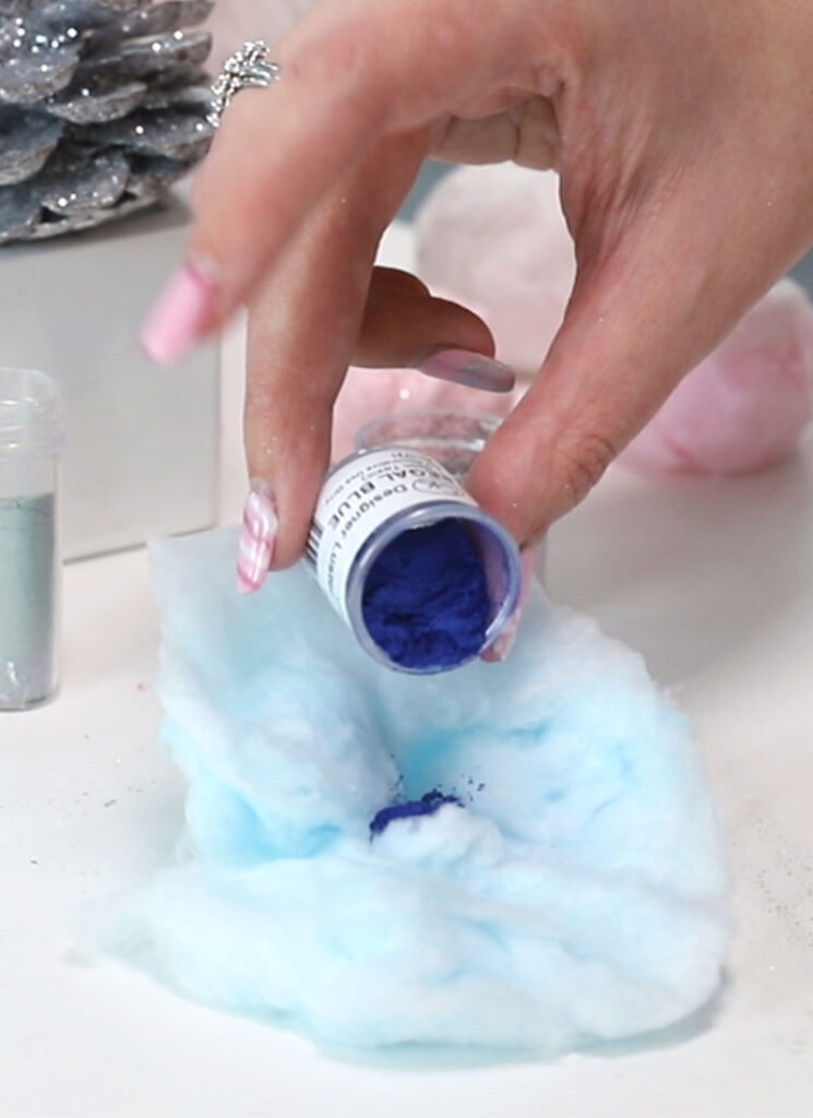 A hand is sprinkling blue glitter from a small jar onto a bed of blue cotton candy.