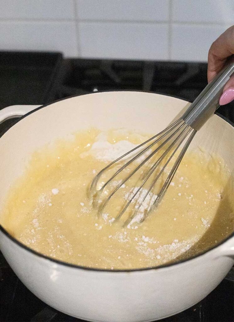 A hand is whisking a large, white pot filled with melted white chocolate fudge. The pot is sitting on a gas stove.