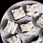 A white plate filled with brownies decorated to look like mummies. The plate is on a black table.