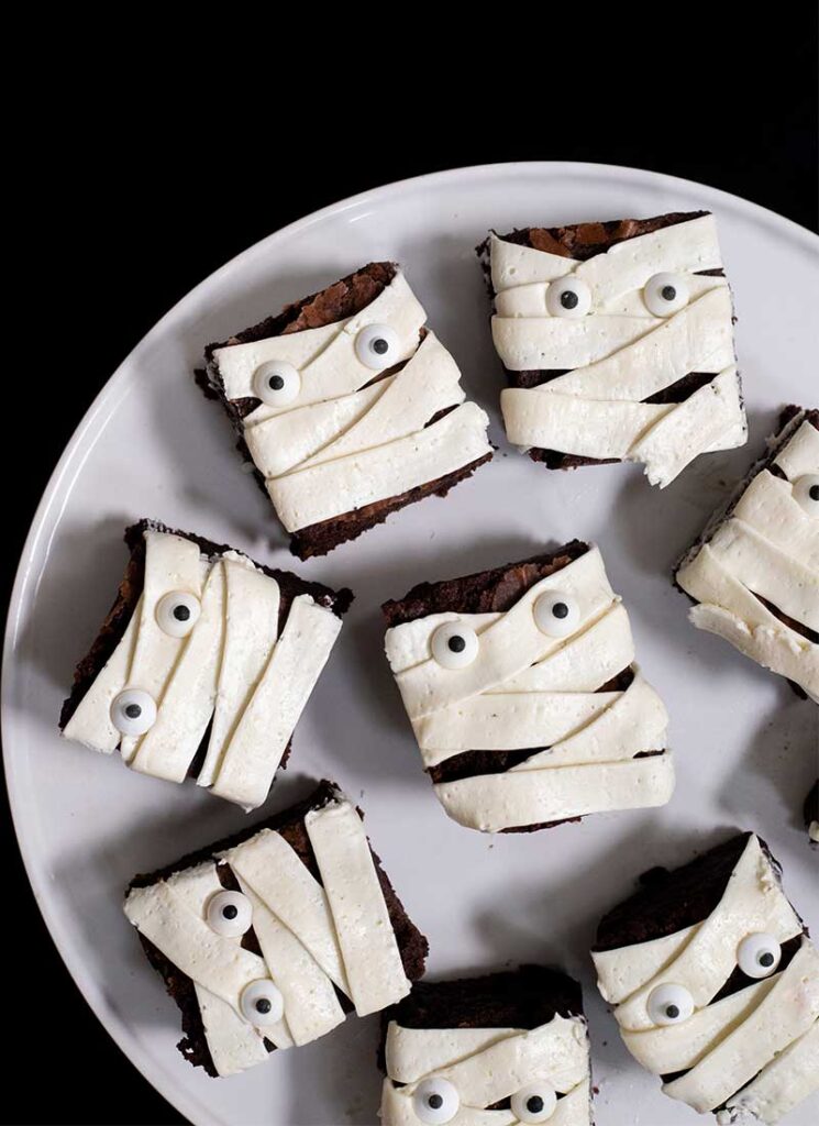 A white plate filled with brownies decorated to look like mummies. The plate is on a black table.
