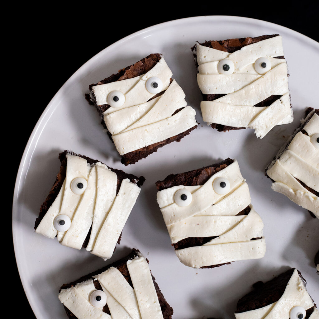 A white plate filled with brownies decorated to look like mummies. The plate is on a black table.