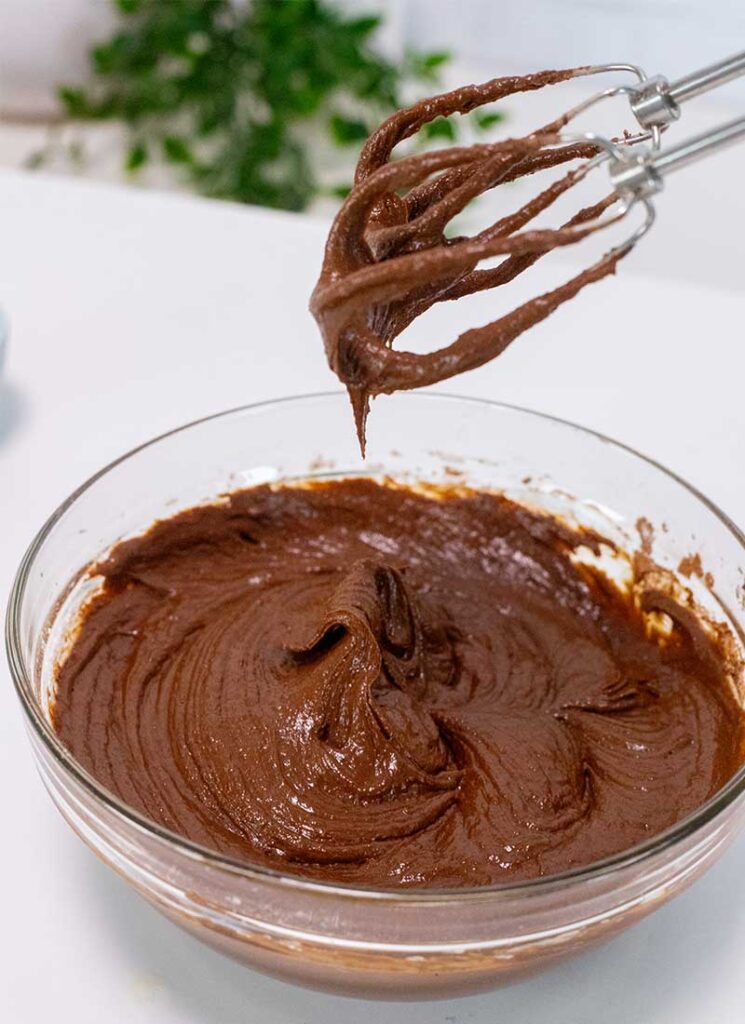 Wet brownie batter is in a large glass bowl. The whisks of a hand mixer are above the bowl, coated in brownie batter.