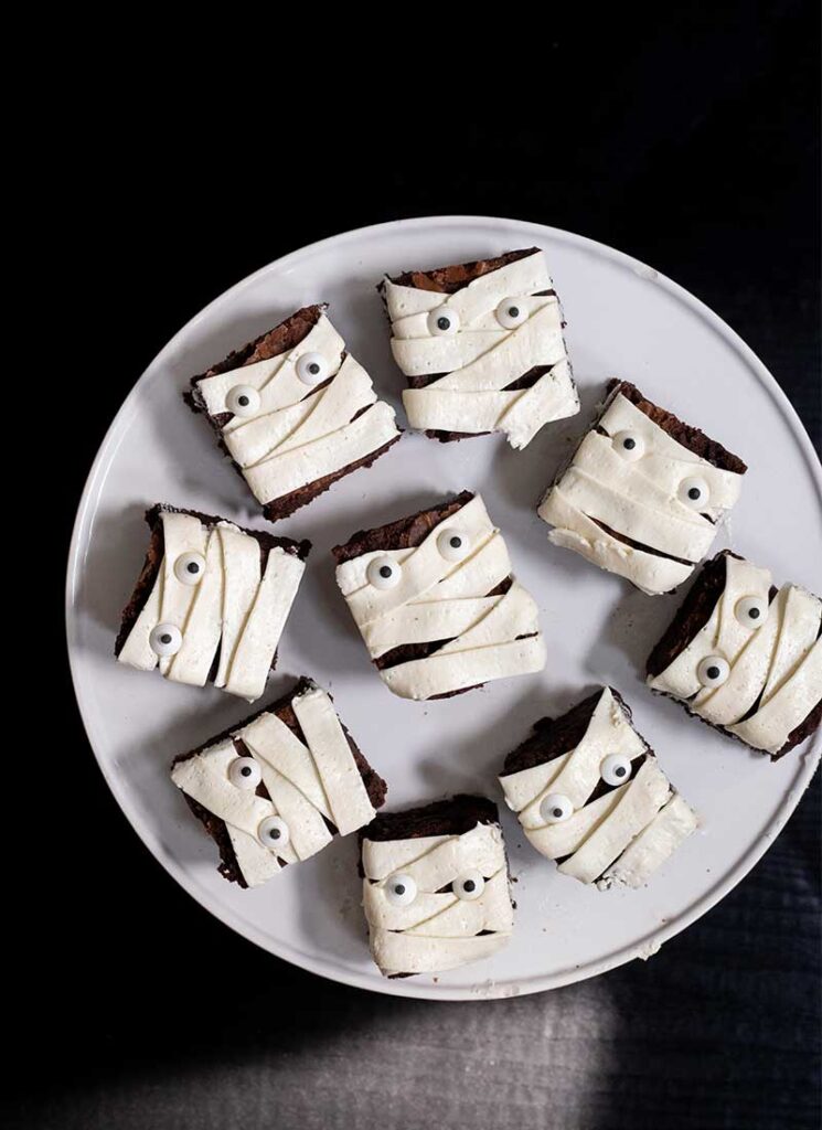 A white plate filled with brownies decorated to look like mummies. The plate is on a black table.