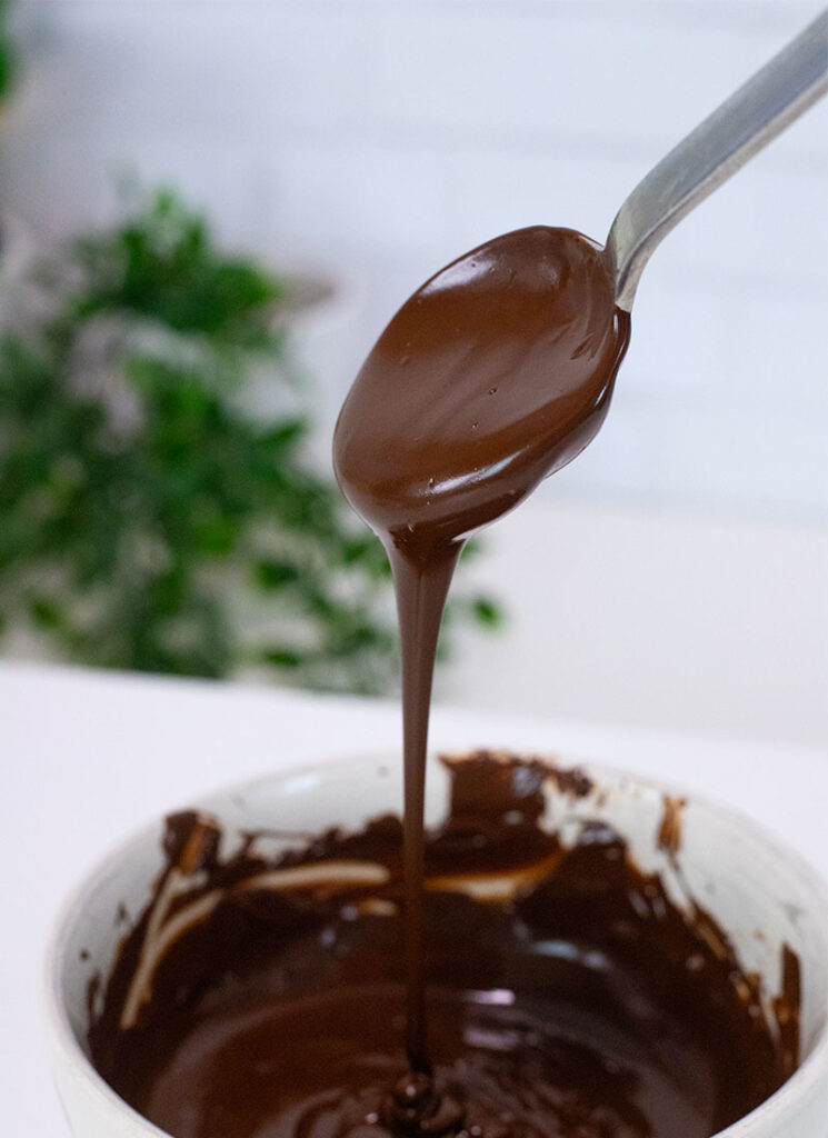 Melted dark chocolate is dripping from a spoon into a white bowl.