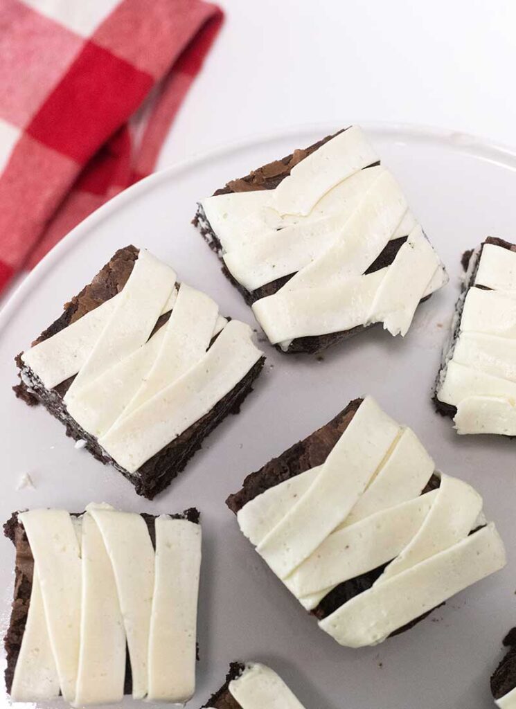 Brownie squares have been decorated with white buttercream to look like mummy bandages. They are on a white plate with a red towel in the background.