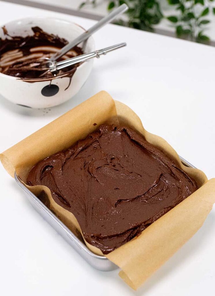 A baked brownie is sitting in its square baking dish that has been lined with brown parchment paper.