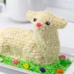 A fully decorated lamb cake sits on a platter, surrounded by green coconut grass and jelly beans.