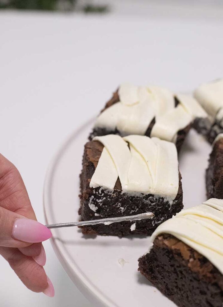 A skewer is scraping off excess frosting on the sides of the brownies.