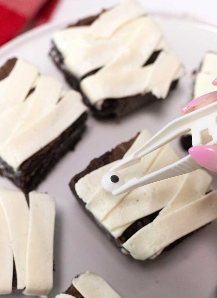 A pair of tweezers are placing a candy eye onto the brownies.