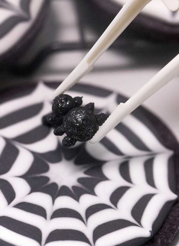White tweezers are placing a sparkly black spider onto a cookie.