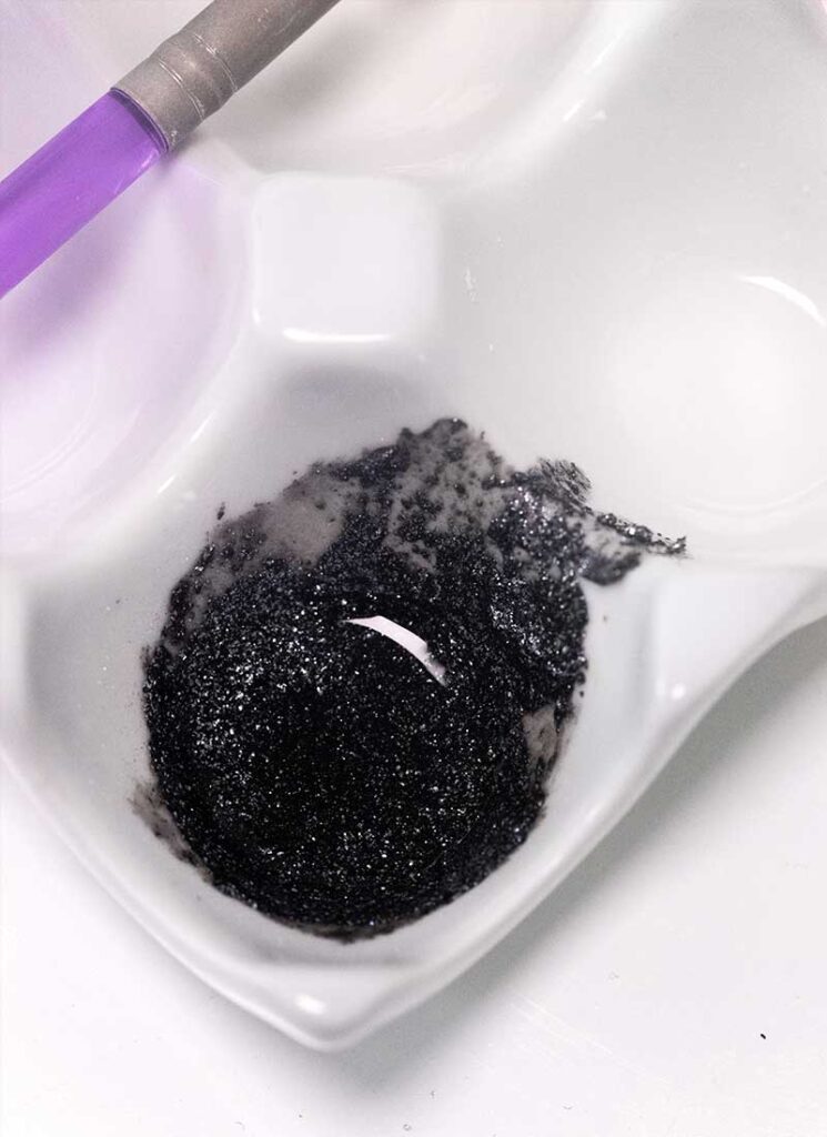 A ceramic dish containing sparkly black liquid edible glitter. A purple brush is off to the side.