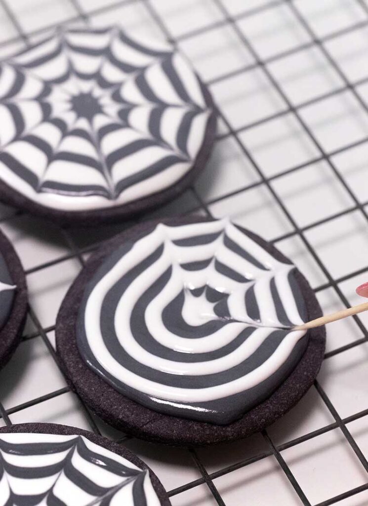 A toothpick is being pulled through the icing on the cookie, creating a spider web-like effect with the white circles.
