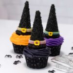 3 witch hat cupcakes are on a white table. The cupcakes are black, purple and orange. Spiders and a skeleton hand are next to the cupcakes.