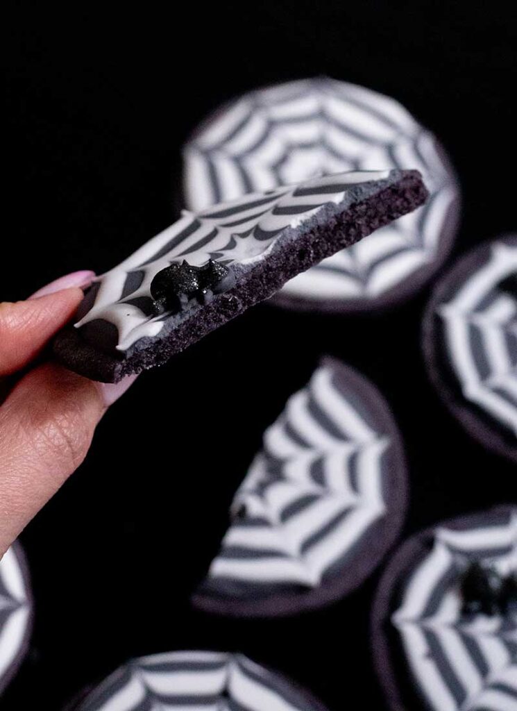 A cookie has been broken in half and is being held up to the camera. You can see the spider and the edge of the cookie. Other cookies are in the background.