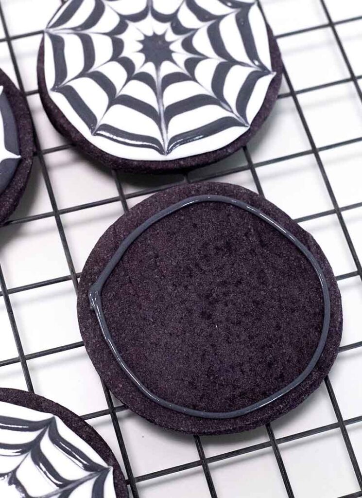A round black cookie is on a wire rack. A ring of black icing has been piped around the edges of the cookie. The cookie is surrounded by other fully decorated cookies.