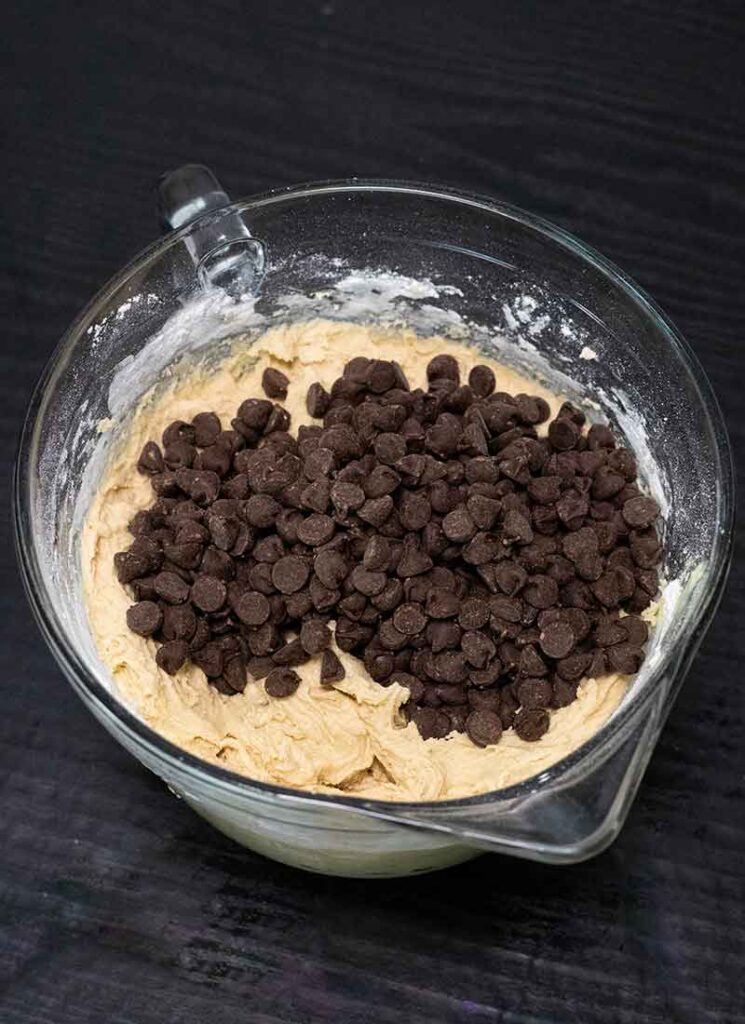 Chocolate chips have been poured into a large glass mixing bowl containing cookie dough. It has not been mixed in yet.