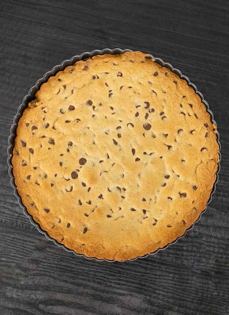 The giant cookie has been baked and is cooling in its pan. It is on a black table.