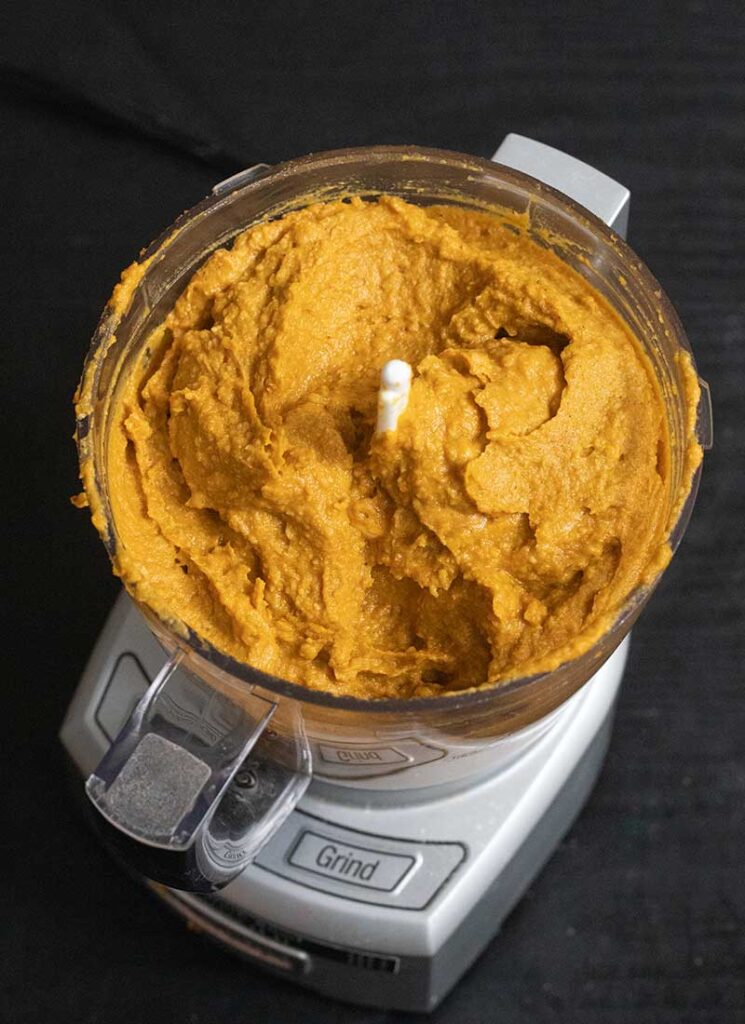 The blended pumpkin hummus is in a blender with the lid taken off. It is a vibrant orange color and looks smooth and creamy.