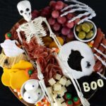 A Halloween-themed charcuterie board, featuring a whole plastic skeleton, with meats, fruit and cheeses surrounding it.
