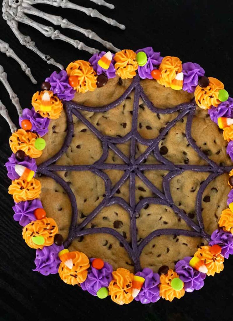 A top down view of the Halloween cookie cake. It is on a black table with skeleton hands reaching for the cookie from the top left.