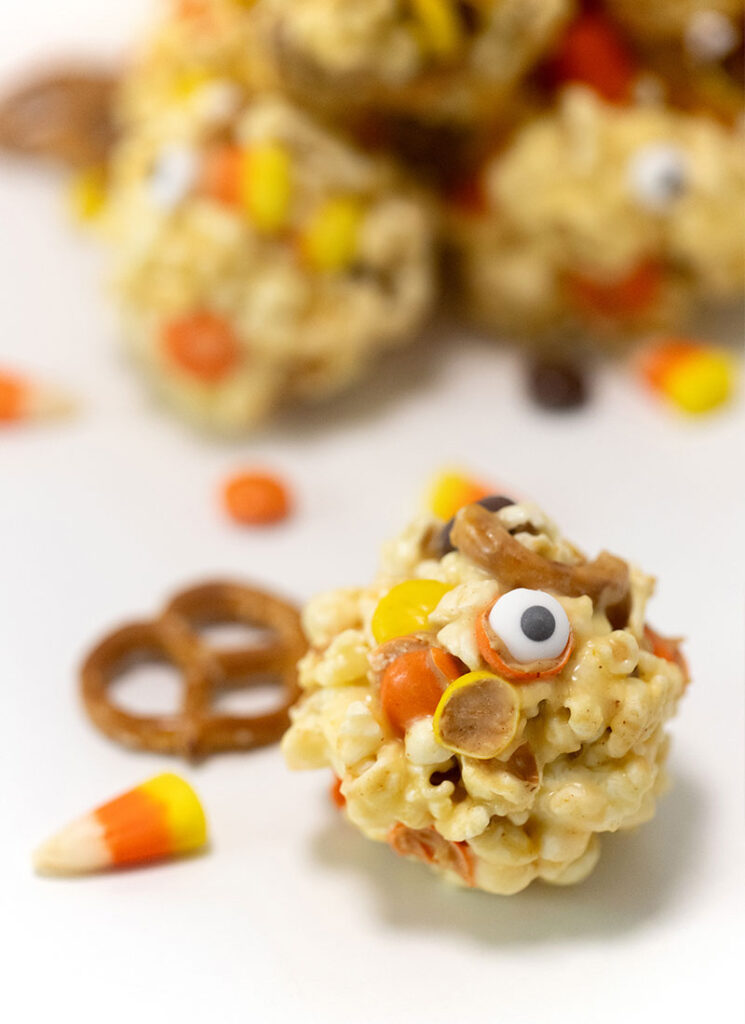 A Halloween popcorn ball is sitting in the foreground next to a pretzel and piece of candy corn. More popcorn balls are in a pile in the background.