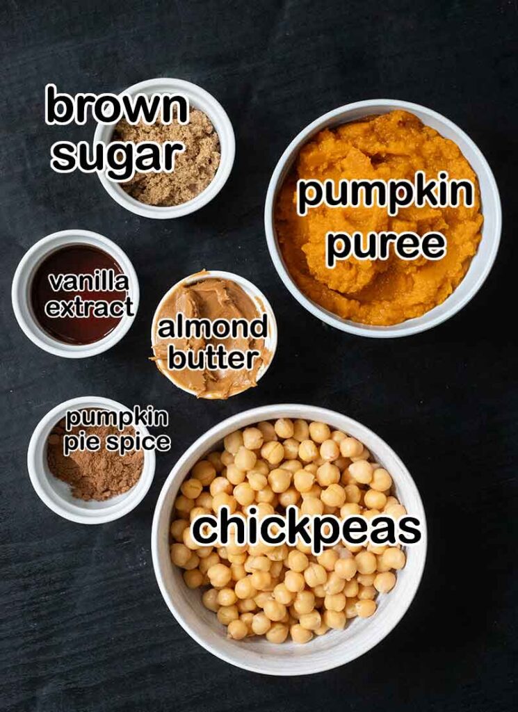 A top down view of the ingredients for pumpkin pie hummus. They are arranged on a black table and are in white bowls.