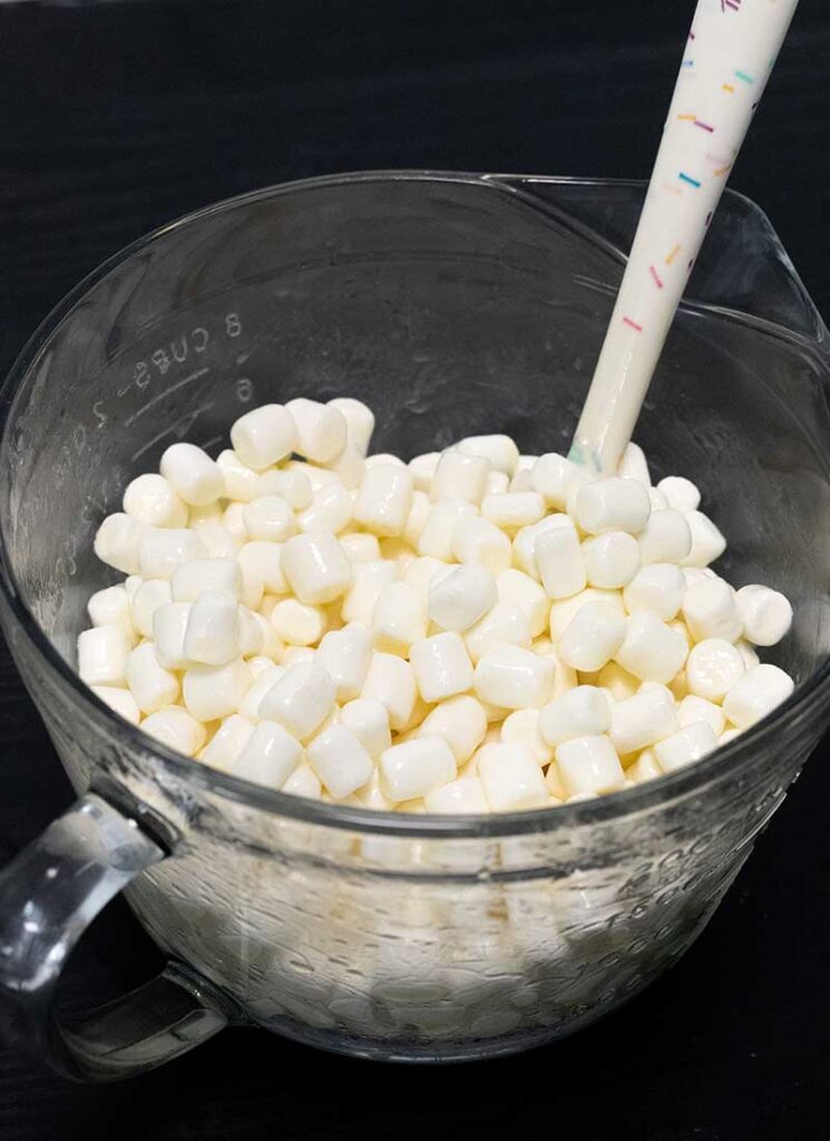 A large glass mixing bowl contains mini marshmallows that have been coated in melted butter. A white spatula is also inside the bowl.