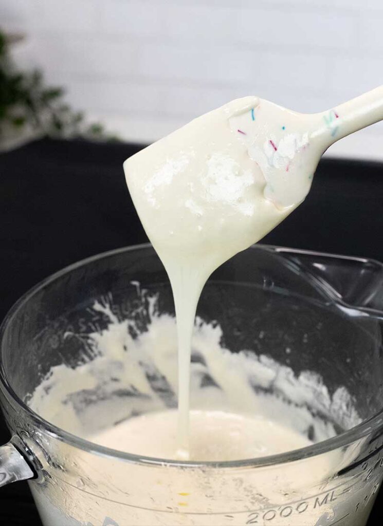 Melted marshmallows are dripping from a white spatula into a large glass mixing bowl.