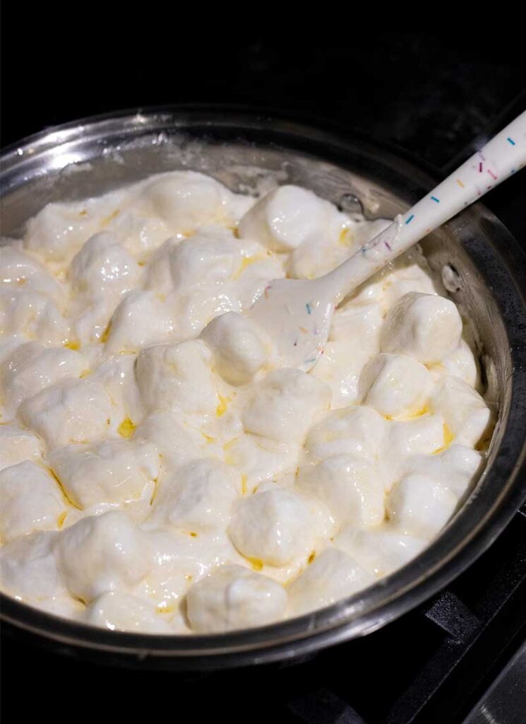 A pot filled with jumbo marshmallows. They are beginning to melt into the butter. A white spatula is about to stir them.