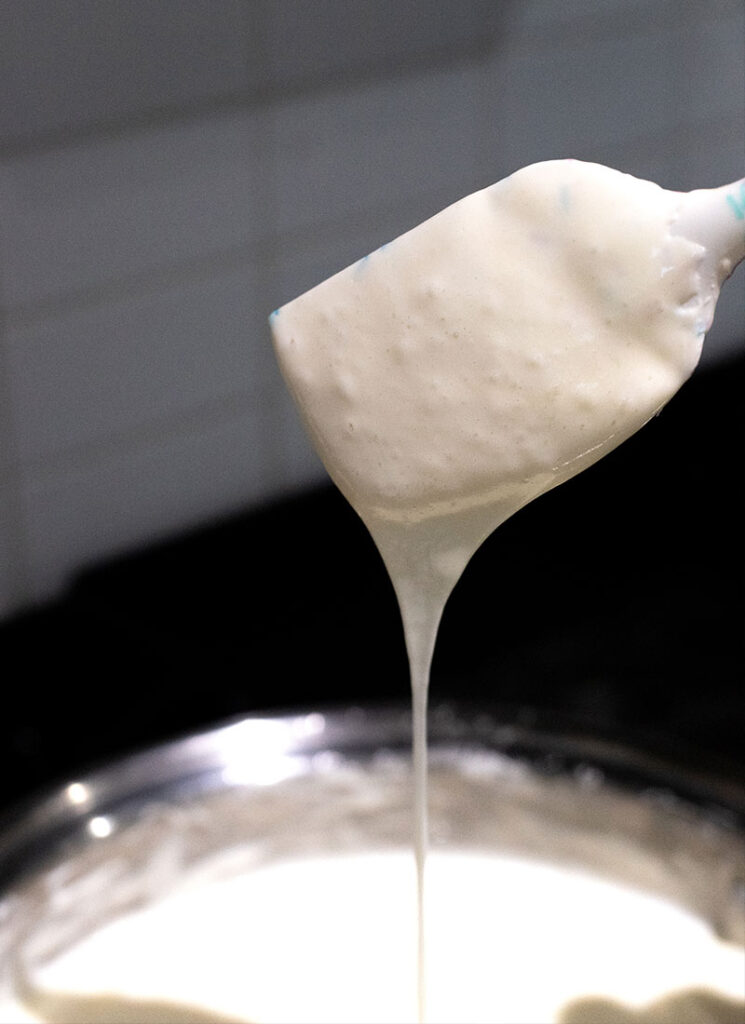 Melted marshmallow is dripping off a white spatula into a pot filled with more marshmallow.