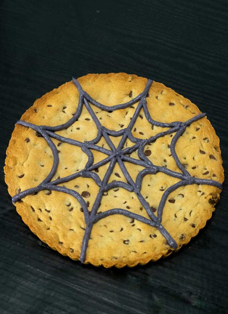 A large black buttercream spiderweb has been piped onto the surface of the giant cookie.