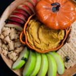 A wooden tray filled with graham crackers, teddy grahams and sliced red and green apples. A ceramic pumpkin-shaped dish is in the middle and is filled with pumpkin pie hummus. The lid to the dish is slightly off to the side. Small black plastic spiders are scattered over the tray.