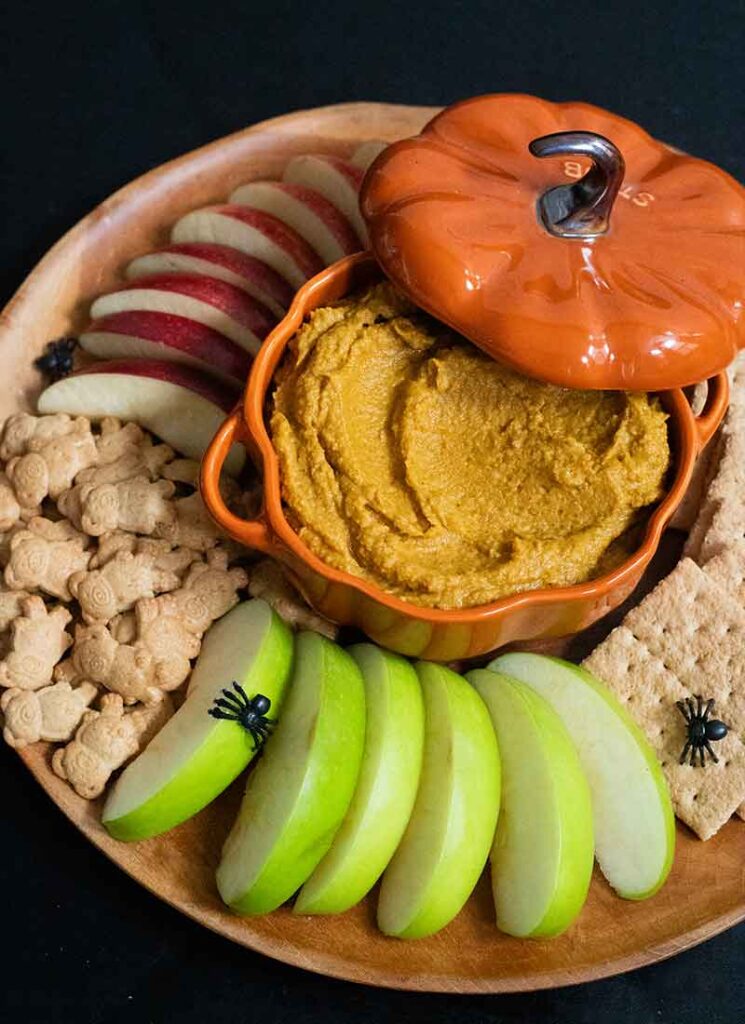 A wooden tray filled with graham crackers, teddy grahams and sliced red and green apples. A ceramic pumpkin-shaped dish is in the middle and is filled with pumpkin pie hummus. The lid to the dish is slightly off to the side. Small black plastic spiders are scattered over the tray.