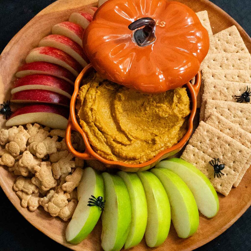 A wooden tray filled with graham crackers, teddy grahams and sliced red and green apples. A ceramic pumpkin-shaped dish is in the middle and is filled with pumpkin pie hummus. The lid to the dish is slightly off to the side. Small black plastic spiders are scattered over the tray.