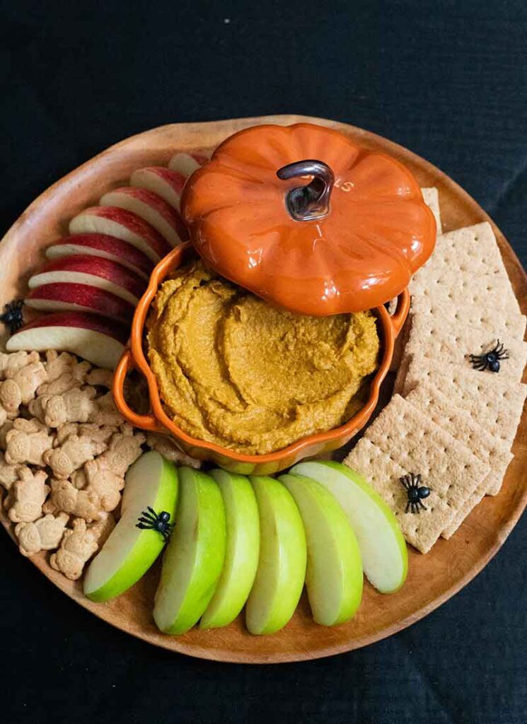 A wooden tray filled with graham crackers, teddy grahams and sliced red and green apples. A ceramic pumpkin-shaped dish is in the middle and is filled with pumpkin pie hummus. The lid to the dish is slightly off to the side. Small black plastic spiders are scattered over the tray.