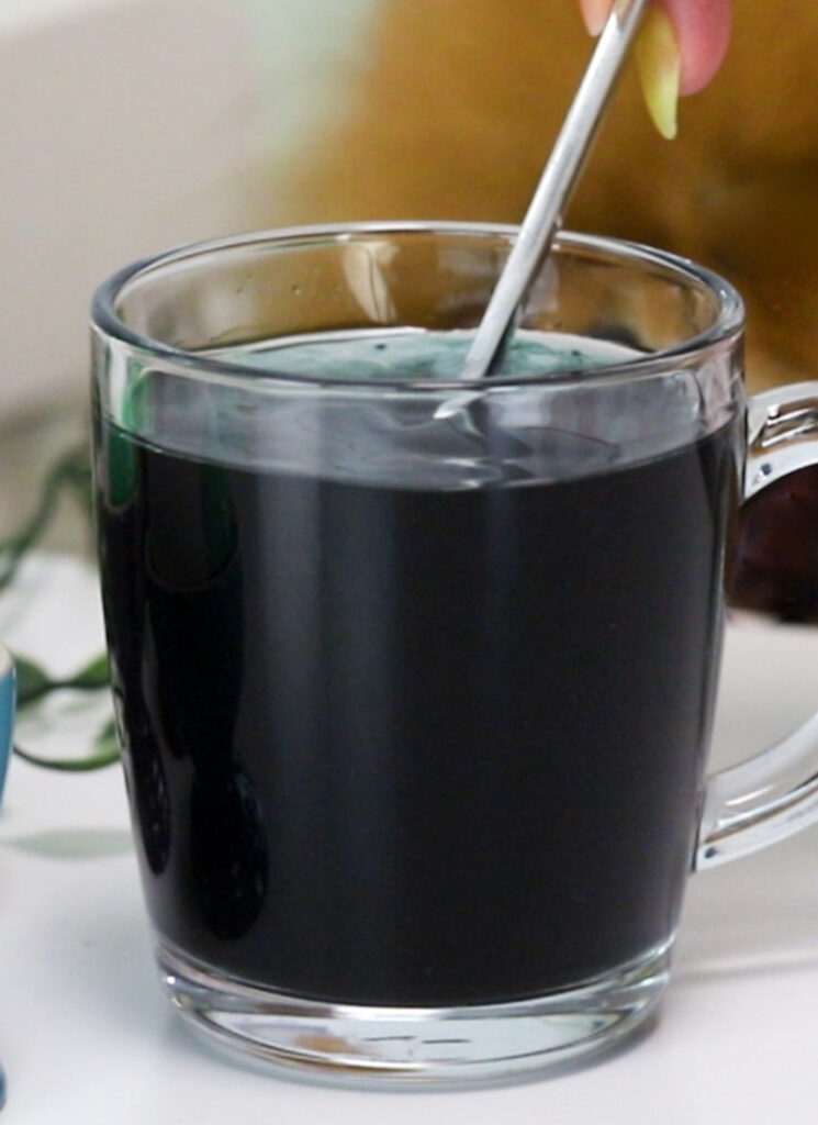 A spoon is stirring a glass containing water, vinegar and black food coloring.
