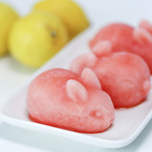 Three rabbit-shaped pink lemonade drink bombs sit on a white plate. Lemons are in the background.