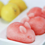 Three rabbit-shaped pink lemonade drink bombs sit on a white plate. Lemons are in the background.