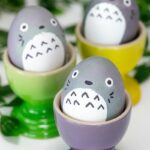 3 Easter eggs decorated to look like Totoro are sitting in purple, green and yellow eggs cups. A green plant is behind them.